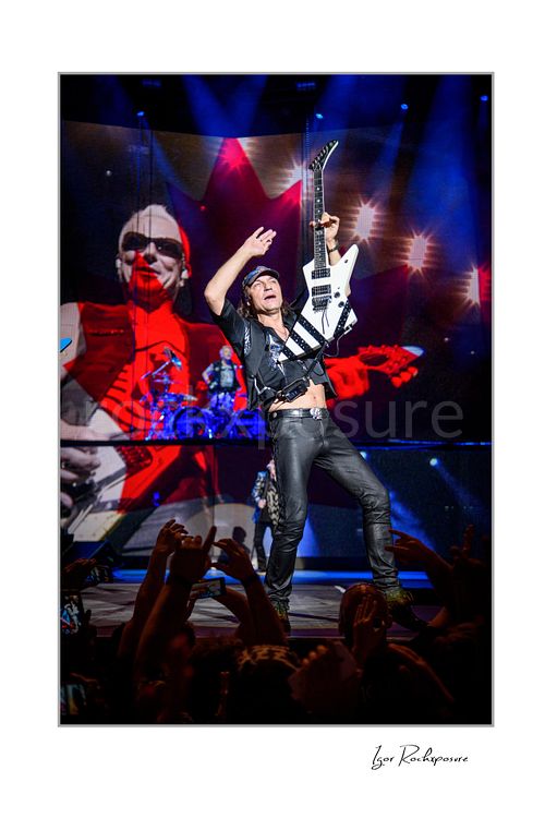 Vertical color image of Rudolf Schenker of Scorpions performing live with white guitar raised, crowd hands in foreground, and giant screen backdrop
