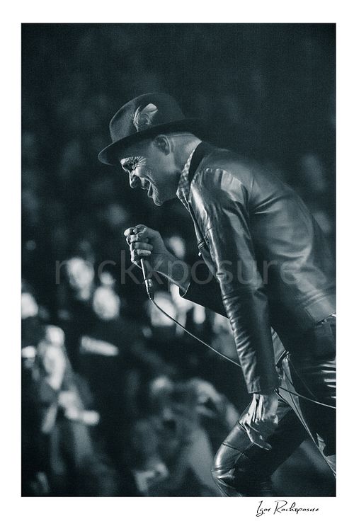 Vertical black and white image of Gord Downie of The Tragically Hip leaning forward with microphone in hand during a live performance, wearing a hat and leather jacket with the crowd softly blurred behind him