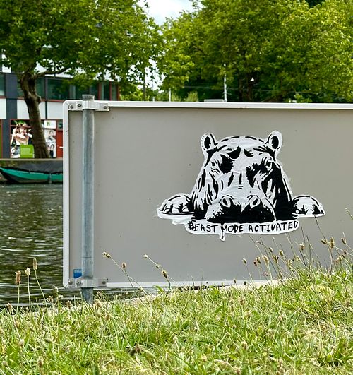 Street art stencil of a hippo's head emerging, with the text "#BEAST MODE ACTIVATED" below it. Located on a sign next to a canal in Amsterdam, this piece by Street Cyber symbolizes the raw, unstoppable power and resilience of decentralized movements, hinting at the awakening of individual financial strength through Bitcoin.