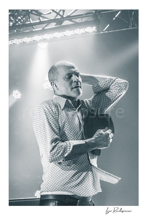 Vertical black and white image of Gord Downie of The Tragically Hip in an emotional live performance moment, holding his hat to his chest with one hand behind his neck under soft stage light