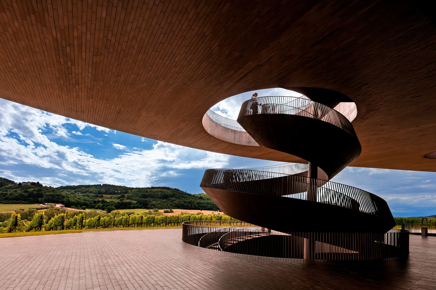 Low-angle architectural view of the weathered Cor-Ten steel spiral staircase at the Antinori winery in Tuscany, ascending through a circular opening in a terracotta-clad ceiling toward a blue sky.