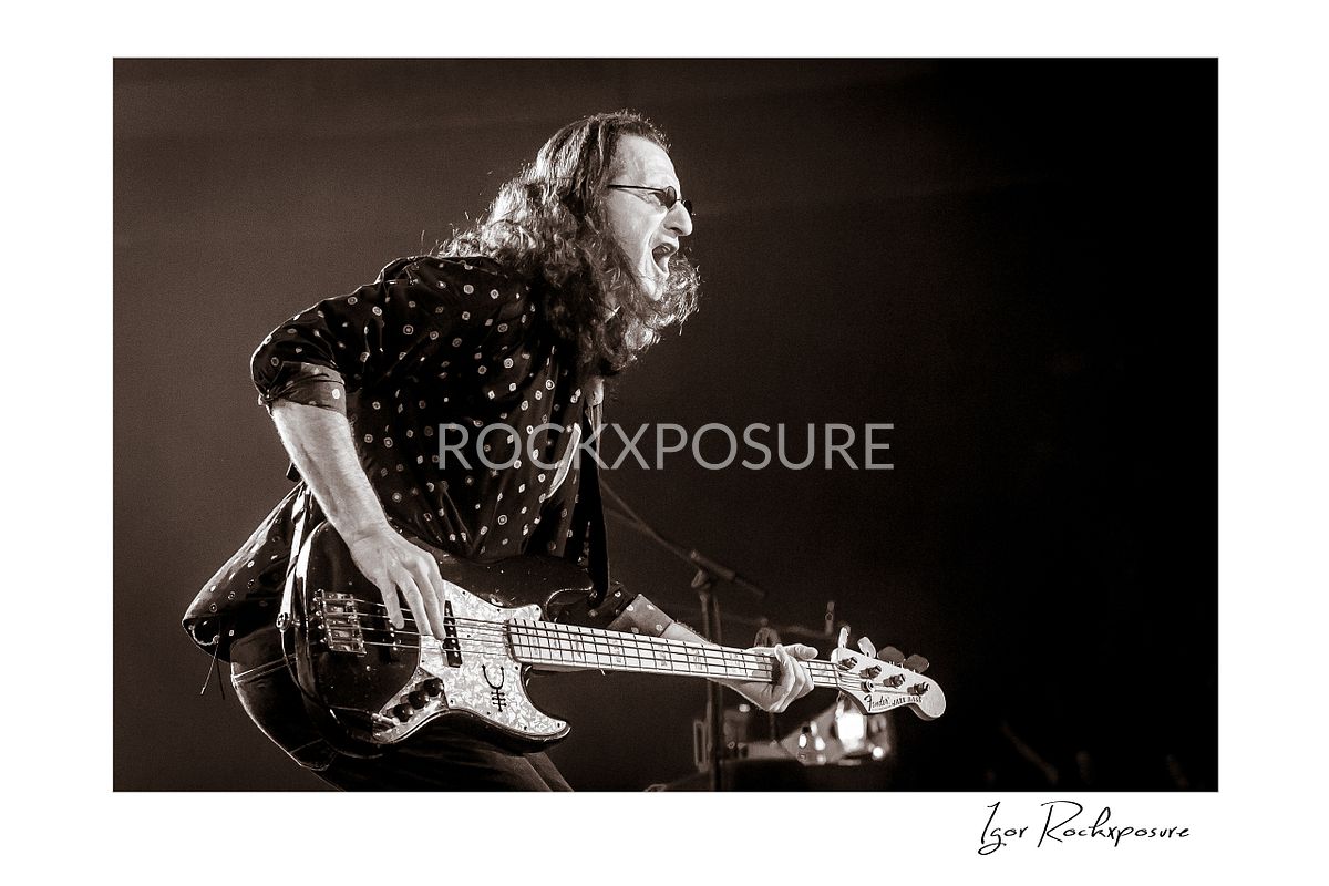 Horizontal concert photography of Geddy Lee leaning forward while playing bass guitar under stage lighting