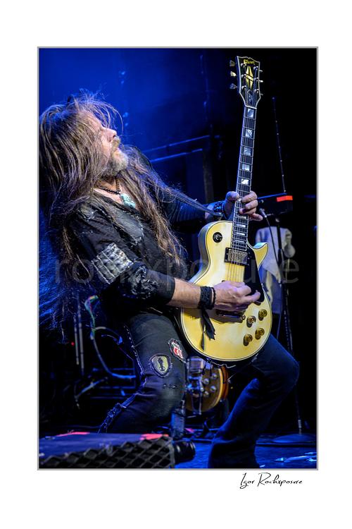 Vertical color image of Chris Latham of Beasto Blanco playing guitar under vivid blue stage light, with long hair in motion and a cream Les Paul style guitar
