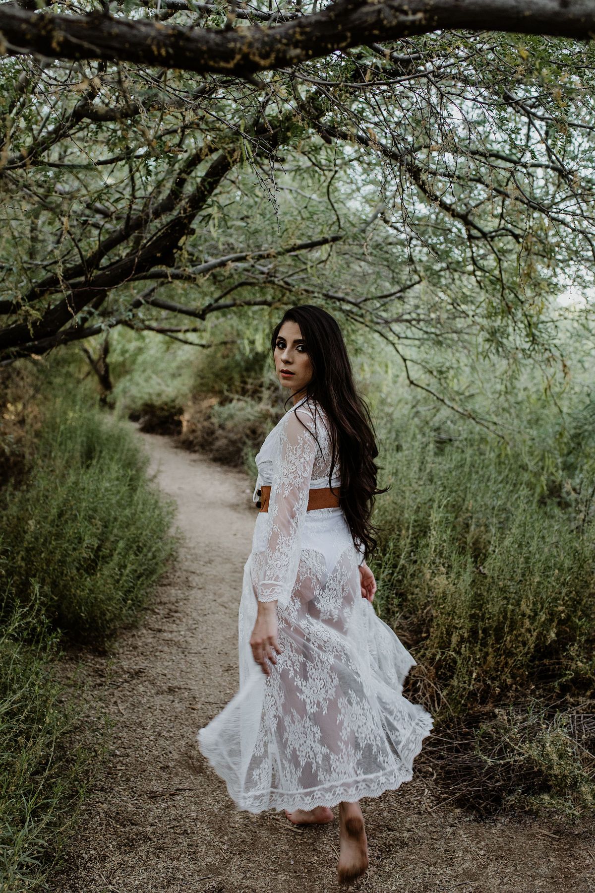 Dreamy boudoir photo of a woman running barefoot and looking back with a smile, wearing a white lace robe in a lush, green tree tunnel — full of movement, freedom, and natural beauty.