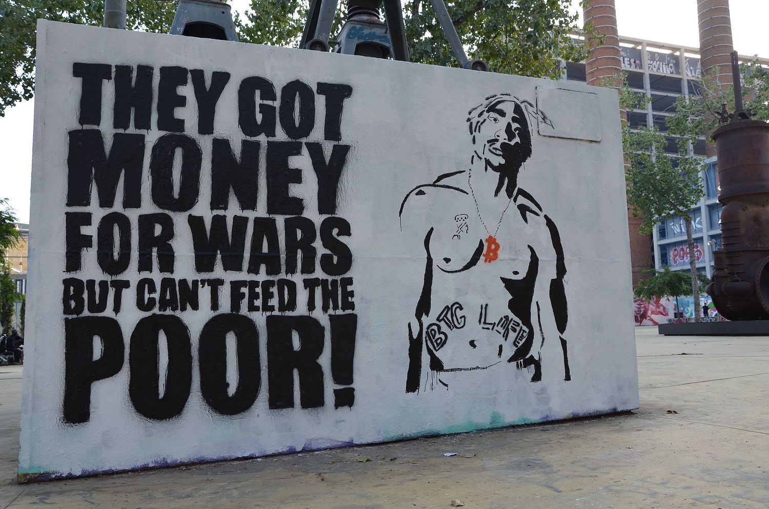Street art mural by Street Cyber featuring a black stencil of Tupac Shakur alongside the bold text "THEY GOT MONEY FOR WARS BUT CAN'T FEED THE POOR!". Tupac has an orange Bitcoin symbol necklace and a "BTC LIFE" tattoo. This powerful piece critiques societal financial priorities, advocating for economic justice and highlights Bitcoin's role in a more equitable world. It blends pop culture with strong social commentary.
