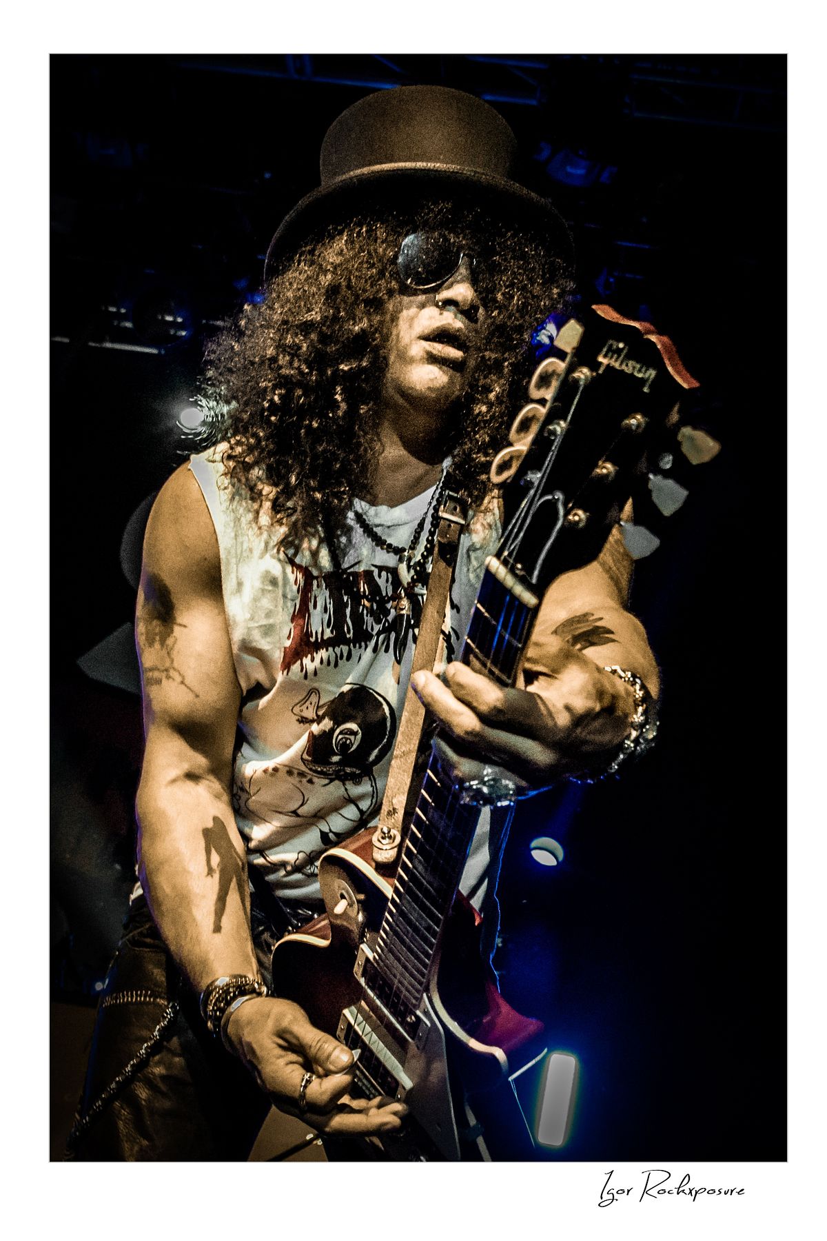 Vertical concert photography of Slash playing electric guitar wearing his signature top hat and sunglasses under dramatic stage lighting