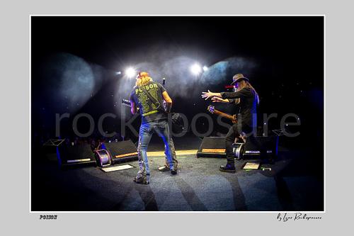 Horizontal color image of Bret Michaels and C.C. DeVille on a Poison stage, photographed with a remote camera mounted on the drum kit facing forward, with bright backlights and stage smoke