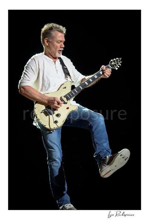 Vertical color image of Eddie Van Halen of Van Halen performing live with a white electric guitar and one leg raised against a dark stage background