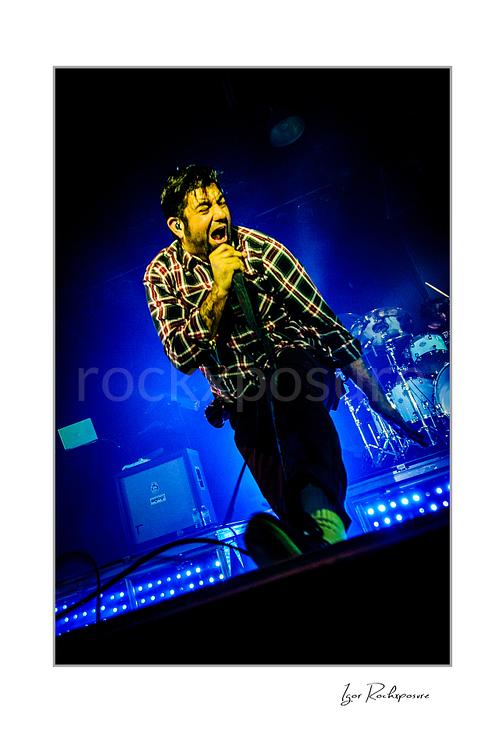 Vertical color image of Chino Moreno of Deftones performing live on stage with a handheld microphone, plaid shirt, blue stage lighting, and the drum kit visible behind him