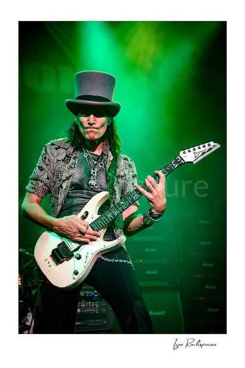 Vertical color image of Steve Vai playing guitar in a tall top hat under vivid green stage lighting