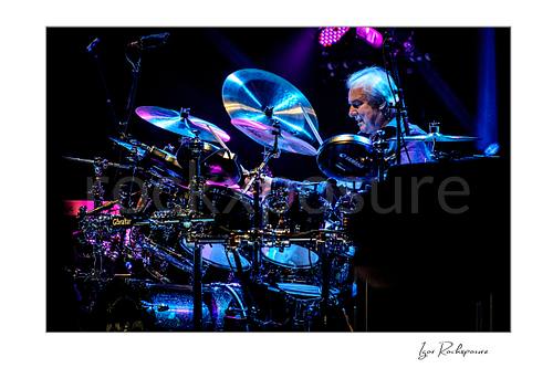 Horizontal color image of Alan White of Yes performing at the drum kit under vivid blue and purple concert lighting