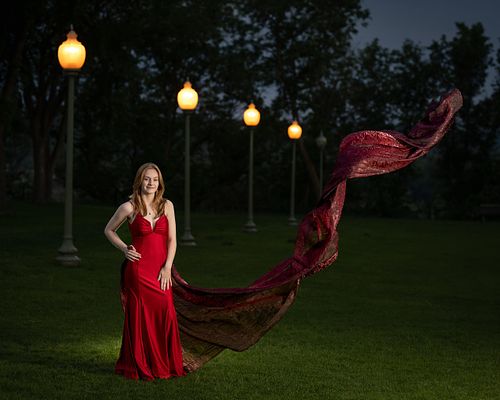 Grad photo of a young woman in a red dress with fabric toss in front of a row of street lights.