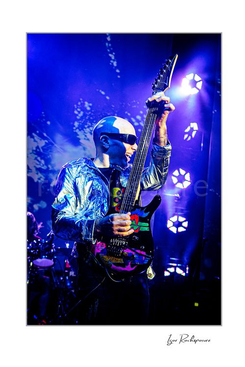 Vertical color image of Joe Satriani performing live in silver jacket and sunglasses with decorated guitar held upright under blue and purple stage lights