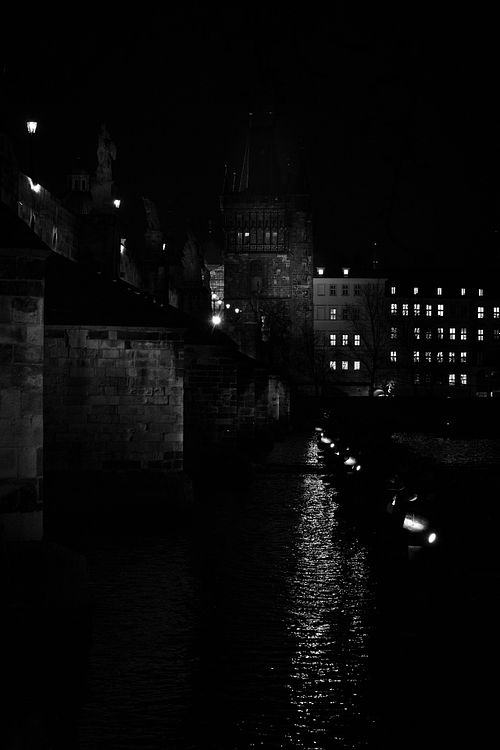 Une vision sombre du pont Charles où la lumière se concentre sur les pierres et les reflets de la Vltava. Une atmosphère dense et silencieuse.