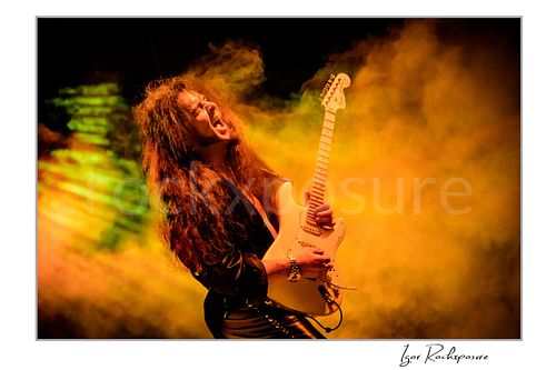 Horizontal color image of Yngwie Malmsteen performing live with a white guitar amid dense golden stage smoke, head thrown back in a dramatic concert moment