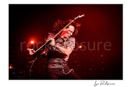 Horizontal color image of Steve Vai swinging his guitar with flying hair under vivid red stage lighting