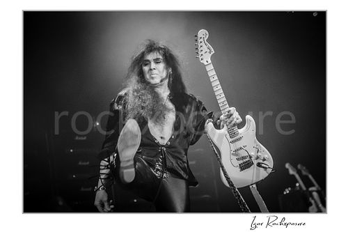 Horizontal black and white image of Yngwie Malmsteen holding a white guitar while delivering another dramatic high kick on stage under soft concert haze