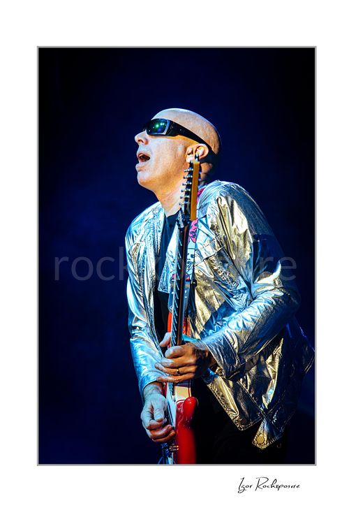 Vertical color image of Joe Satriani performing live in silver jacket and sunglasses with red guitar against deep blue stage light