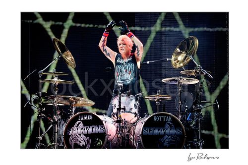 Horizontal color image of James Kottak of Scorpions behind the drums with raised arms, black gloves, and full Scorpions drum setup