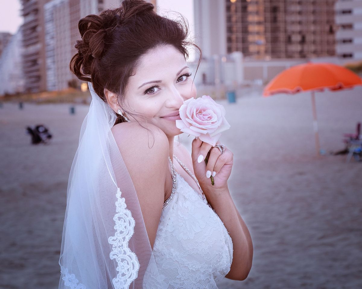 bride is smiling while smelling a pink rose on the beach in sussex county, de