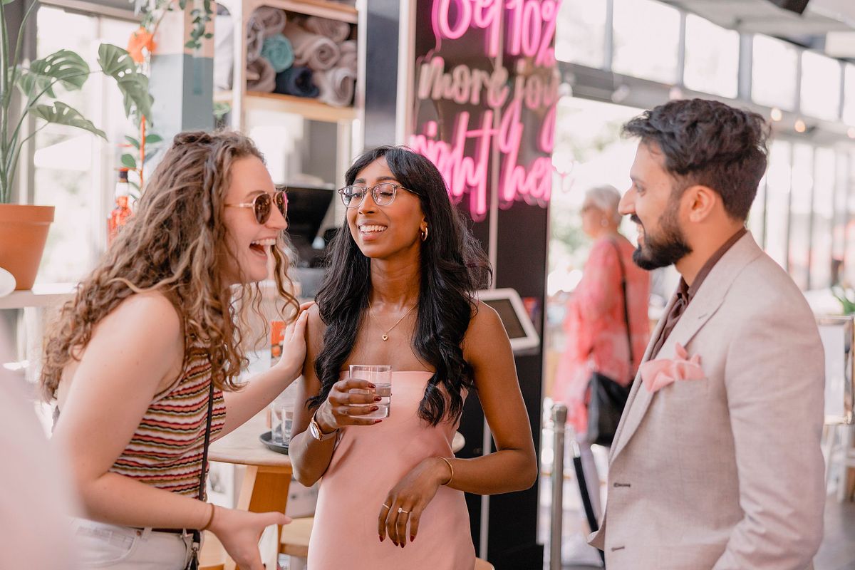Photographer in Basel capturing three friends chatting at an engagement party and laughing in a lively café setting. One holds a drink, with a neon sign in the background, creating a joyful atmosphere.