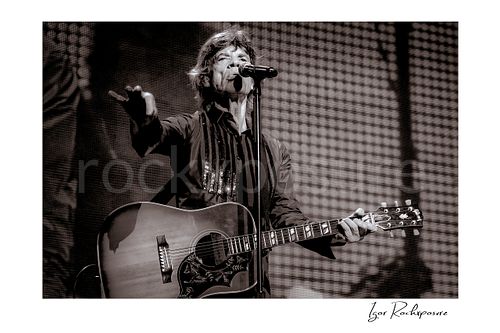 Horizontal black and white image of Mick Jagger of The Rolling Stones singing into a microphone with an acoustic guitar on stage