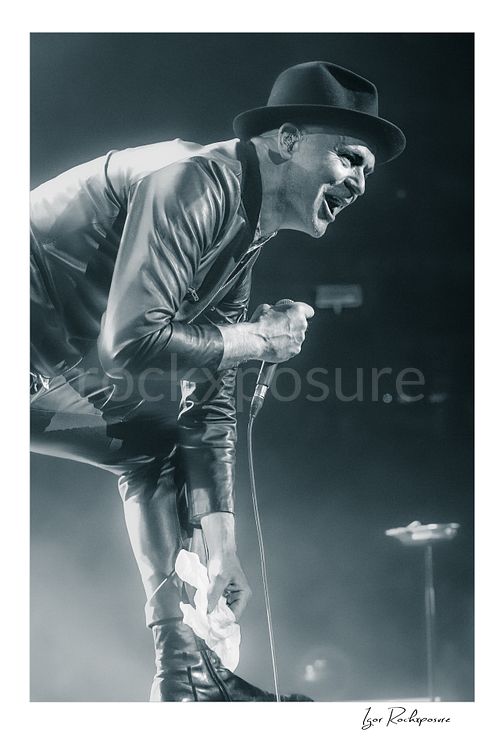 Vertical black and white image of Gord Downie of The Tragically Hip leaning forward and singing into a microphone, wearing a hat and leather jacket with a white cloth in hand amid bright concert haze