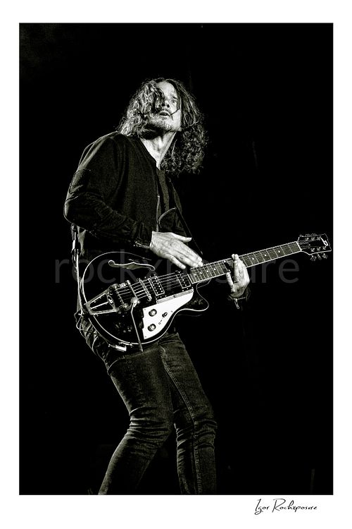Vertical black and white image of Chris Cornell performing live on stage, playing electric guitar against a dark background