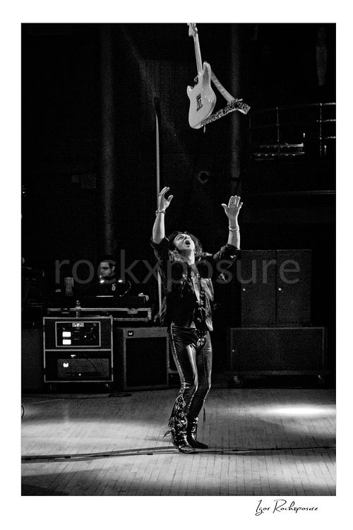 Vertical black and white image of Yngwie Malmsteen tossing his white guitar high above the stage in a dramatic live performance moment, reaching upward beneath it with empty hands