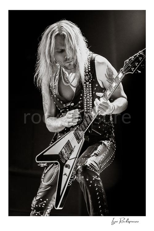 Vertical black and white image of Richie Faulkner of Judas Priest performing live on stage, playing flying V guitar under dramatic concert lighting