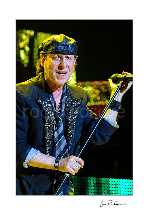 Vertical color image of Klaus Meine of Scorpions singing live with microphone stand, black cap, and sparkling green stage lights