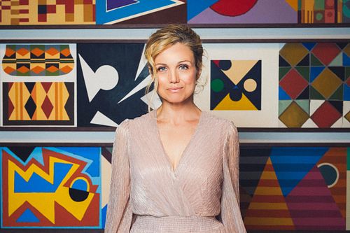 Actress Bridie Carter stands in front of a piece of colourful art with her blonde hair tied back, and wears a beautiful long sleeve silk dress.