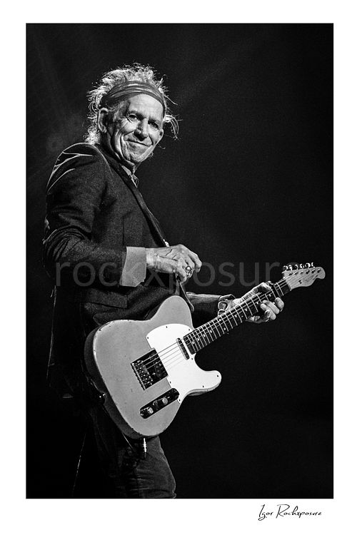 Vertical black and white image of Keith Richards of The Rolling Stones holding an electric guitar in a close-up stage portrait