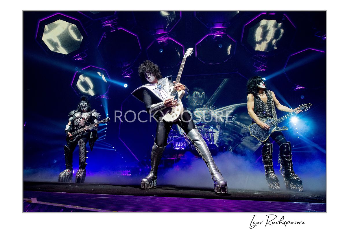 Horizontal color photograph of Gene Simmons, Paul Stanley, and Tommy Thayer performing onstage in full makeup and costumes with purple and blue lighting, hexagonal panels, and the KISS logo visible behind them