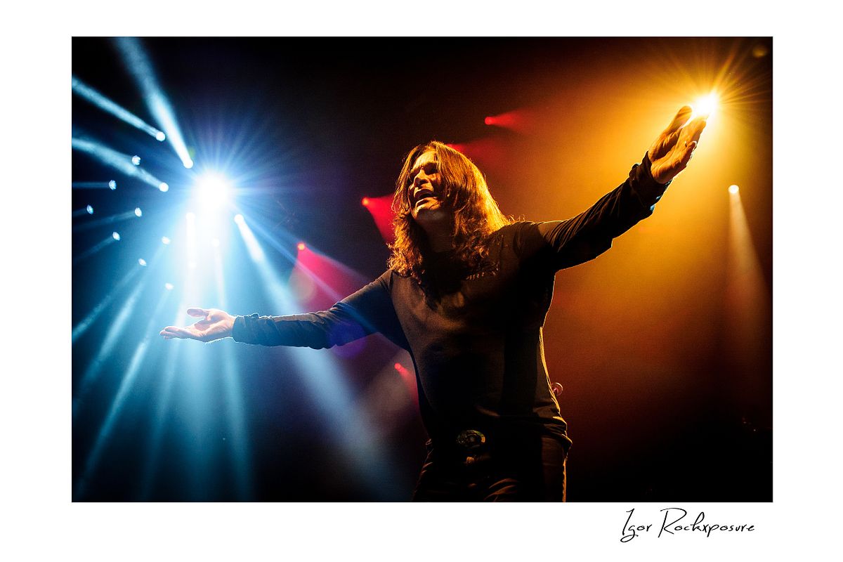 Ozzy Osbourne of Black Sabbath standing with arms raised wide under dramatic blue and orange stage lighting during a live performance