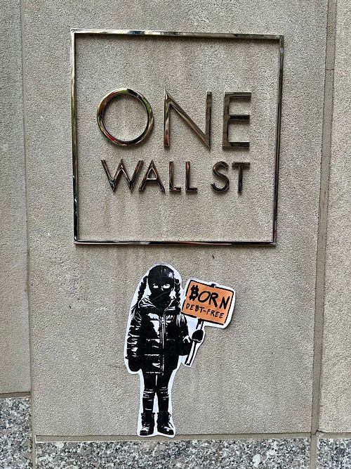 Street art sticker by Street Cyber in New York City, placed directly in front of the "ONE WALL ST" building sign. The sticker depicts a child figure in a black ski mask and jacket, holding an orange sign that reads "BORN DEBT-FREE." This powerful piece directly critiques traditional financial systems and advocates for individual financial freedom and sovereignty through Bitcoin, free from inflationary debt.