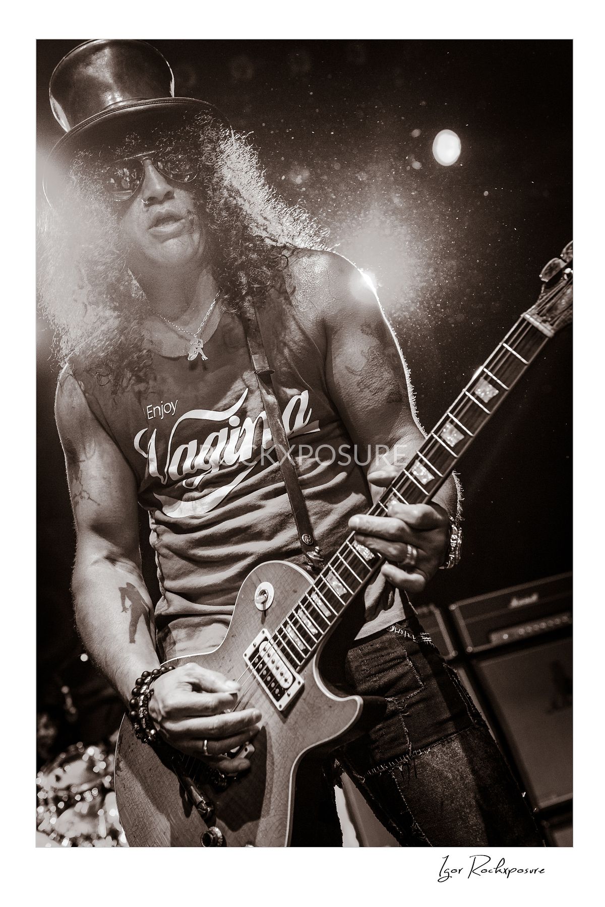 Vertical concert photography of Slash playing electric guitar wearing his signature top hat in black and white concert photography under dramatic stage lighting