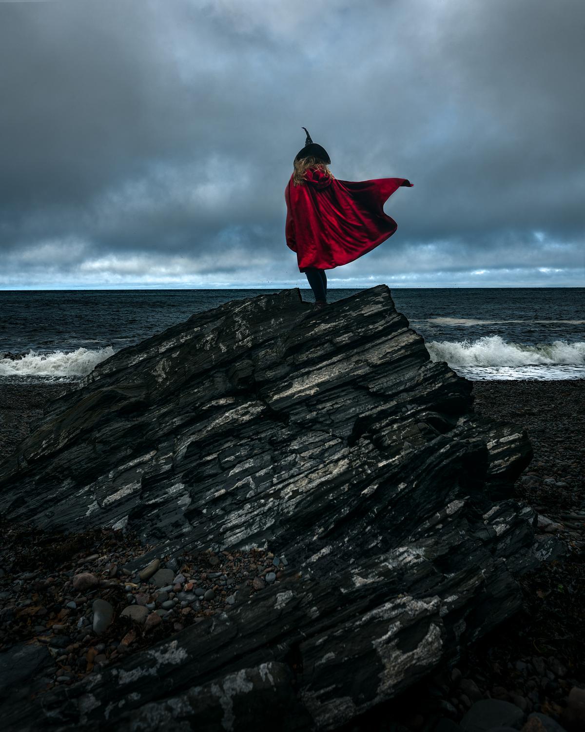 A witch in a red cloak standing on a massive stone looking out to the ocean.