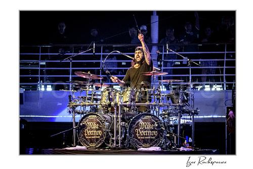 Horizontal color image of Mike Portnoy playing at a double bass drum kit with one drumstick raised high under blue stage lighting