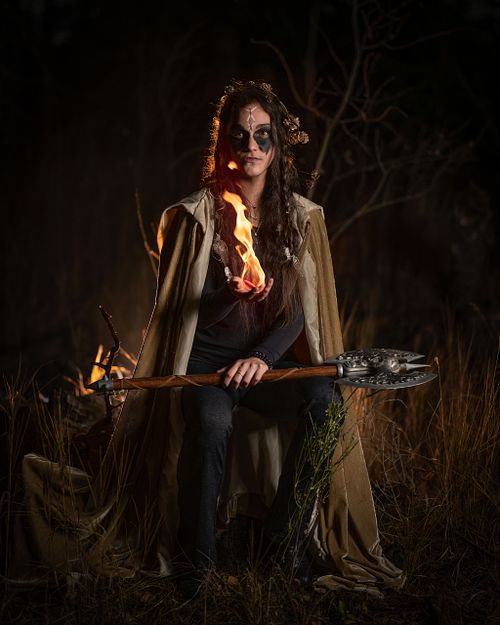 A viking witch wields fire while her battle axe rests on her knees at night.