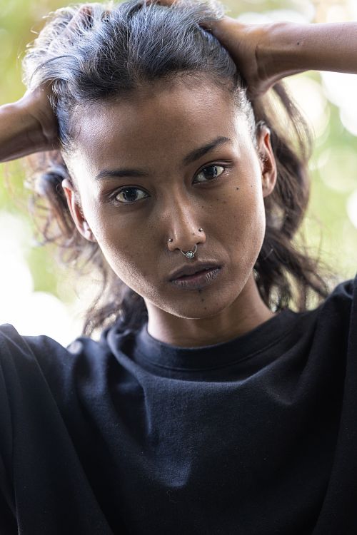 A headshot of an Indian woman, holding her hair with both hands, dressed in a black t-shirt, and adorned with earrings and a nose ring,