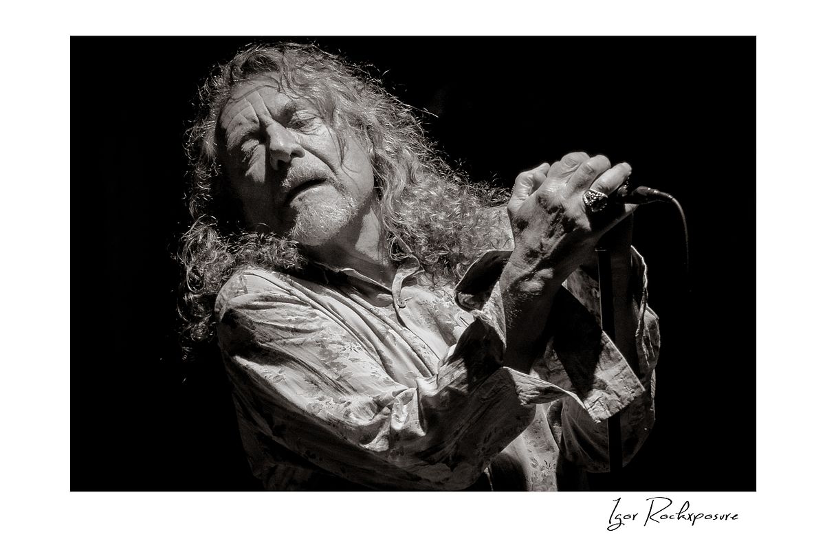 Horizontal black and white concert photography of Robert Plant singing into a microphone with head tilted back during a dramatic live performance
