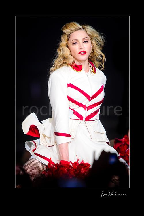 Vertical color image of Madonna in a white and red stage outfit during a theatrical live performance with red pom-poms visible in the foreground