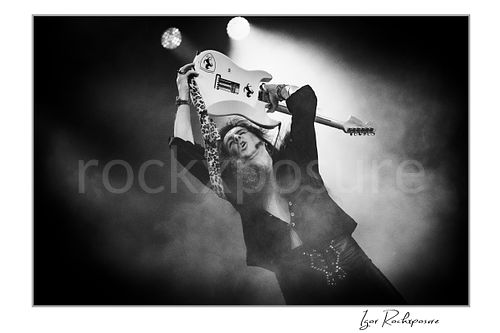 Horizontal black and white image of Yngwie Malmsteen holding a white guitar above his head in a dramatic live performance pose, surrounded by stage smoke and bright backlight