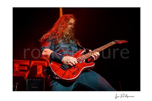 Horizontal color image of Kiko Loureiro of Megadeth playing guitar on stage under red lighting with long hair across his face