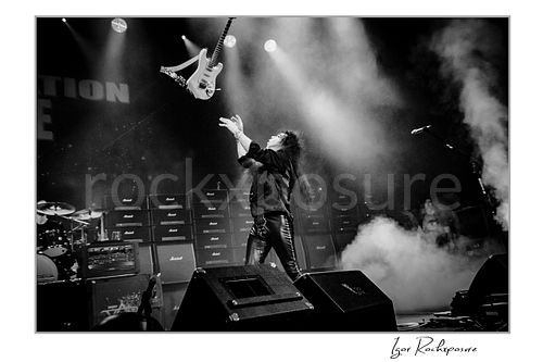 Horizontal black and white image of Yngwie Malmsteen reaching upward to catch his white guitar in mid-air during a dramatic live performance, with stage smoke and a wall of amplifiers behind him